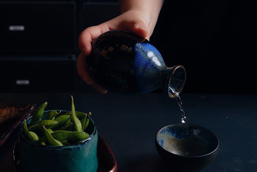 A hand pouring sake from a small blue ceramic bottle into a small ceramic cup, a bowl of edamame beans in the foreground.
