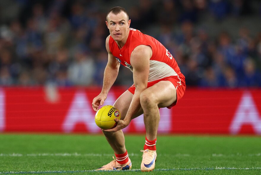 A Sydney Swans AFL player slightly squatting on the field, holding a yellow football in his hand, with blurred fans in the stadium in the background.