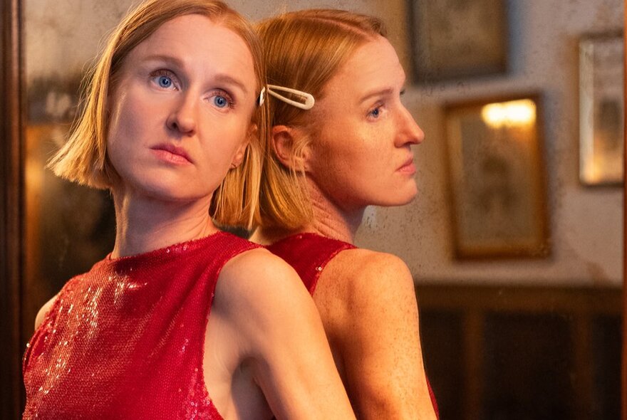 Comedian Nat Harris leaning against a mirror, her reflection showing, looking forlorn and wistful while wearing a sleeveless red dress. 