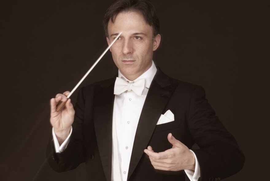 A man wearing a tuxedo and a white bow tie holding a conductor's baton in mid-air, posed against a dark backdrop.