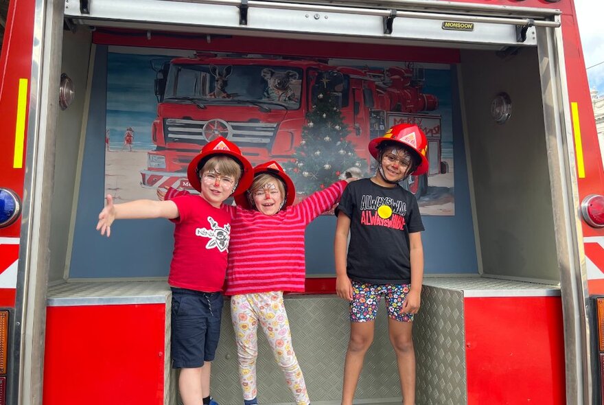 Three children in toy firefighting hats pose in the back of a firetruck.