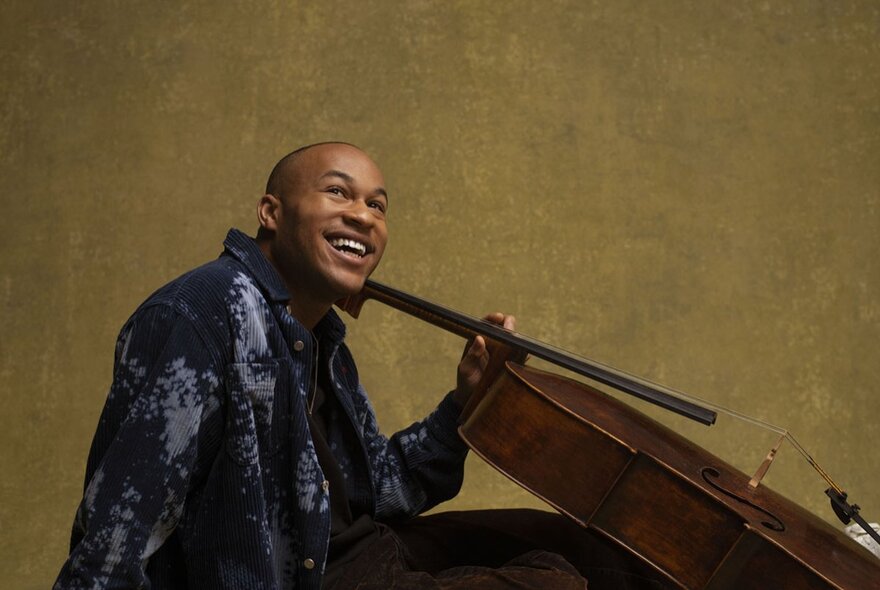 Cellist Sheku Kanneh-Mason smiling and looking upwards, holding the neck of his cello, posed against a wall.