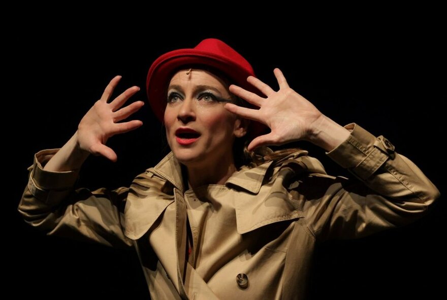 UK performer, Sarah-Louise Young, dressed in a trench coat and red hat, performing in character as Kate Bush on a darkened theatre stage.