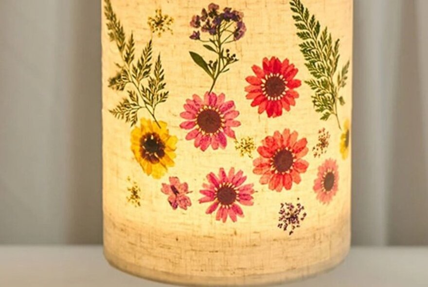 An illuminated lamp with a cylindrical parchment or fabric shade, decorated with various pressed flowers.