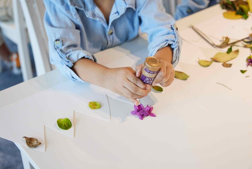 A young child's hands holding a glue stick and attaching leaves and flower petals to paper.