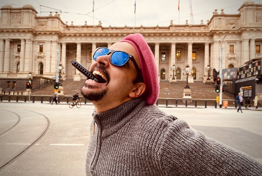 Blind comedian with cigar clamped in his open mouth, wearing glasses, beard and pink beret, on tram tracks in front of Parliament.