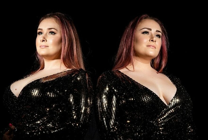 Identical twins standing back to back, wearing matching black dresses with plunging necklines and red hair.