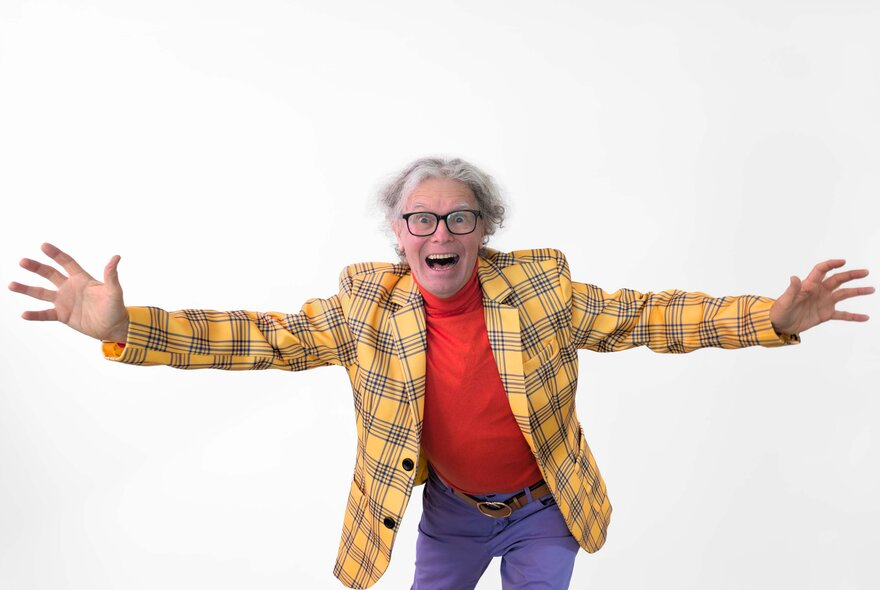 Comedian wearing yellow tartan jacket and red skivvy, with grey hair and spectacles, smiling with arms outstretched.