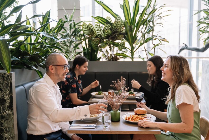 Looking over two tables in a restaurant with potted plants and people in twos chatting over lunch. 
