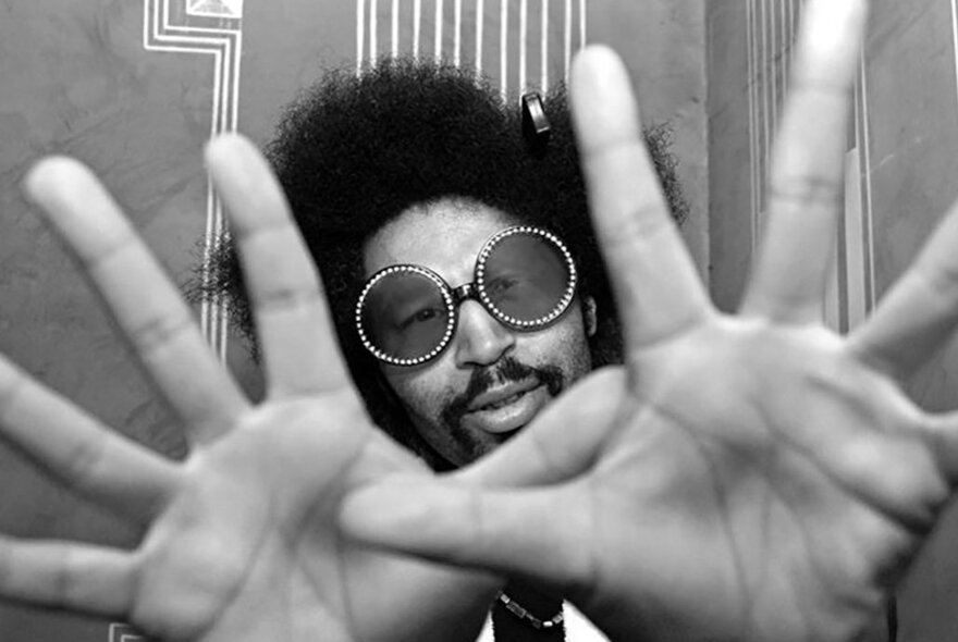 DJ Moodyman, holding his palms stretched out towards the camera, he has curly black hair in an Afro style and is wearing round black sunglasses with a sparkle trim.