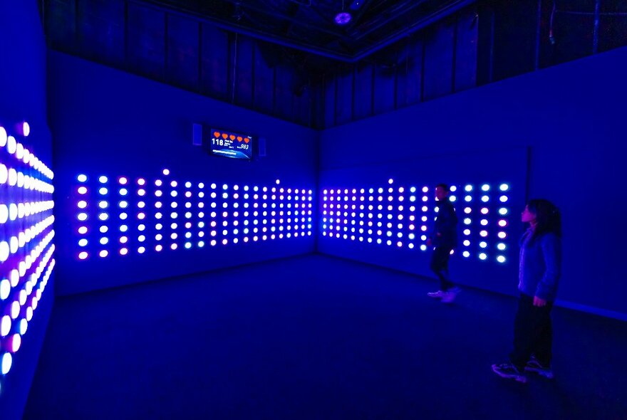 Two people walking into a dark room that has illuminated small circular dome lights embedded in three of the surrounding walls.