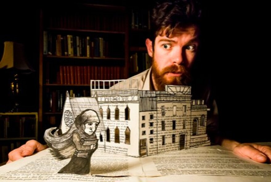A bearded man looking fearfully over the top of a paper model of a streetscape resting on an open book, alongside a drawing of a young woman with a scarf on her head.