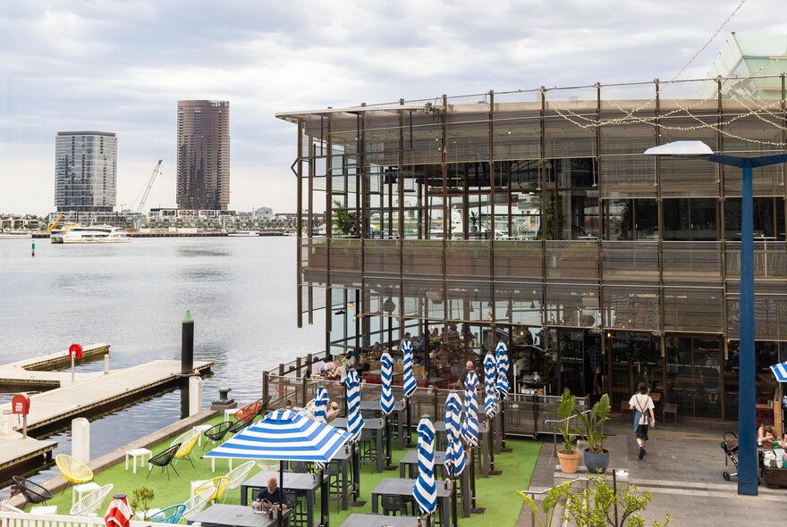 The outdoor seating area of Cargo restaurant, showing blue and white striped shade umbrellas over tables, with a view of the harbour and two large buildings in the distance.
