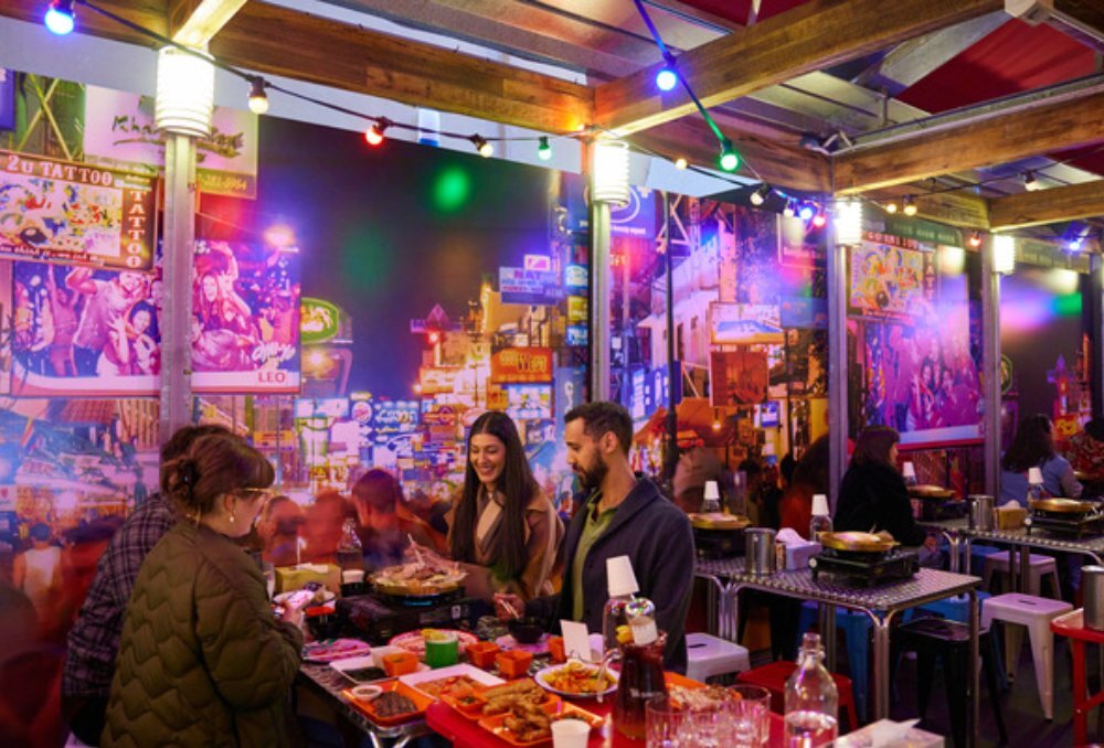 Four friends sitting at a table next to neon art work. The table has a barbecue grill and many small side dishes.