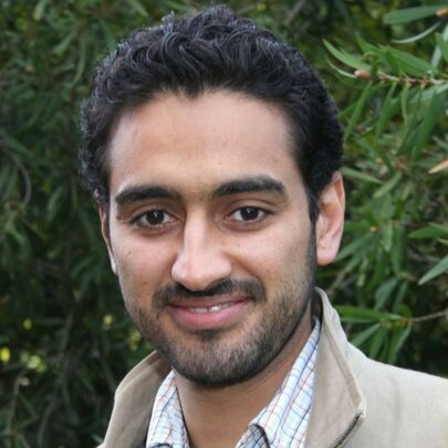 Journalist and academic Waleed Aly, a man with short dark hair and a cropped beard, posed outdoors against a green leafy background.