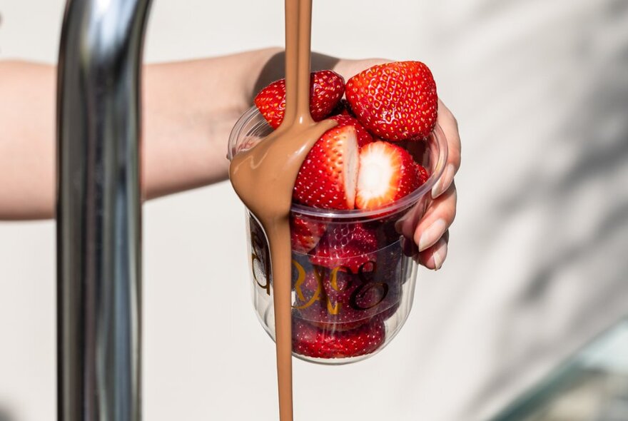 A hand holding a cup of sliced strawberries under a chocolate fountain, the chocolate pouring over the side of the cup.