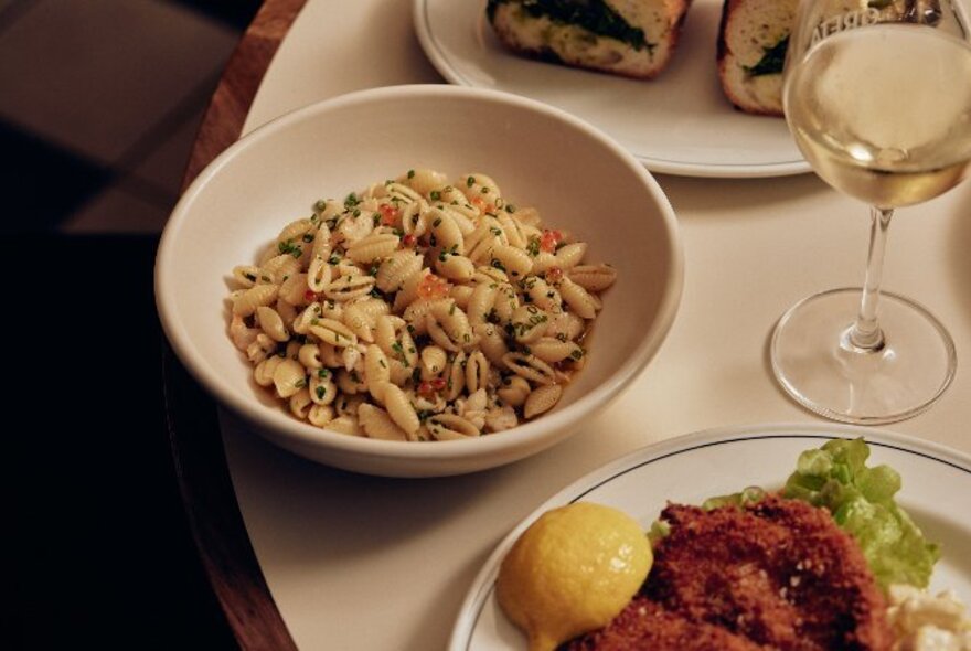 Table shot if a bowl of pasta, glass of white wine and other dishes. 