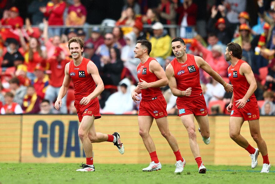 Four Gold Coast Suns AFL players smiling and looking happy on the football field, with a blurred crowd of AFL fans in the stadium behind them. 