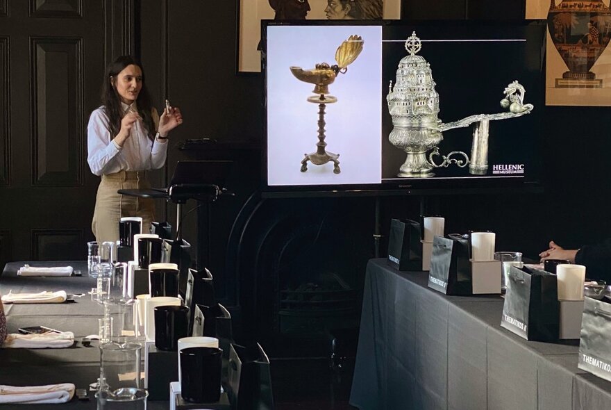 An instructor in front of a large screen talking about scents in a room with tables and chairs ready to host a candle making workshop.