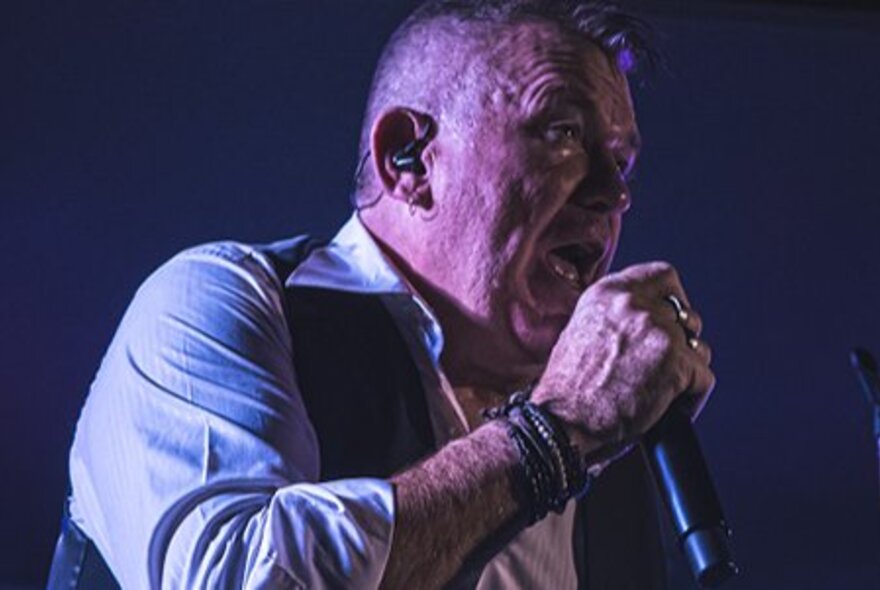 Cold Chisel tribute artist, Steve Cal, singing into a microphone and wearing a vest and white shirt.