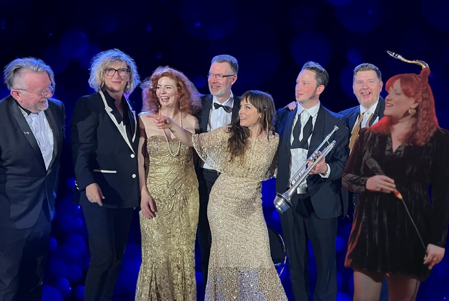 A group of musicians on stage celebrating; three women dressed in evening gowns and the rest of the group in tuxedos.