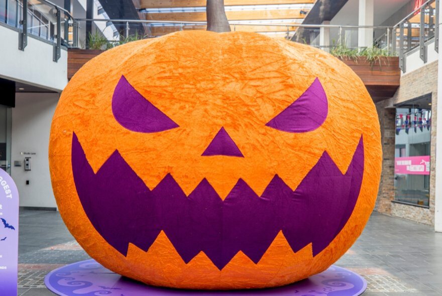 A large, orange inflatable Halloween jack-o'-lantern with a textured surface, a brown stem on top, and sharp, purple facial features of  triangular eyes and a jagged mouth.