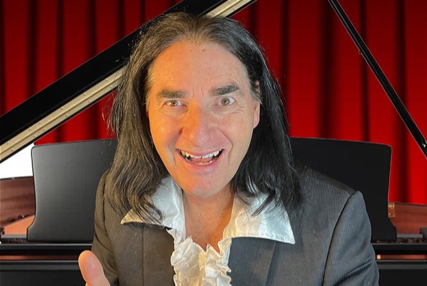 A man with long black hair and frilly shirt smiles intently in front of a piano and red stage curtain. 