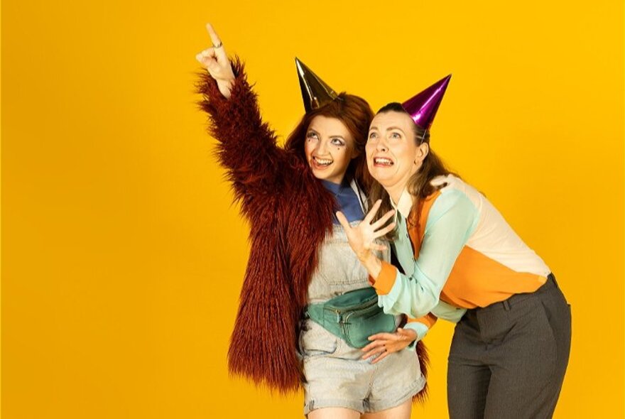 Two women wearing party hats, both looking up at where one of them is pointing - one looking frightened, the other smiling; yellow background.