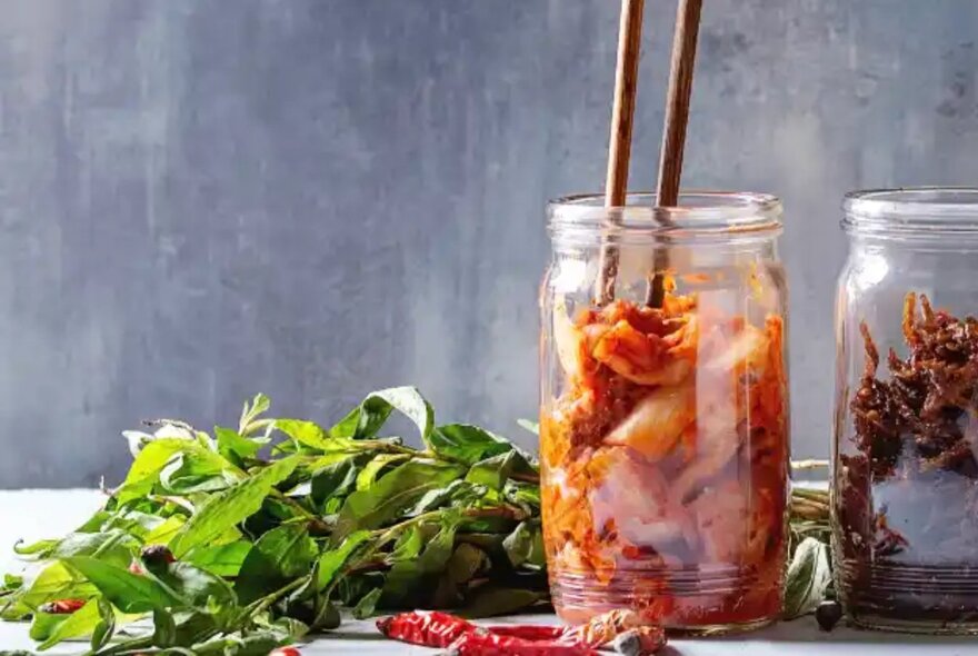 A jar of orange coloured kimchi with two chopsticks poking out of it, and a bunch of fresh herbs alongside.