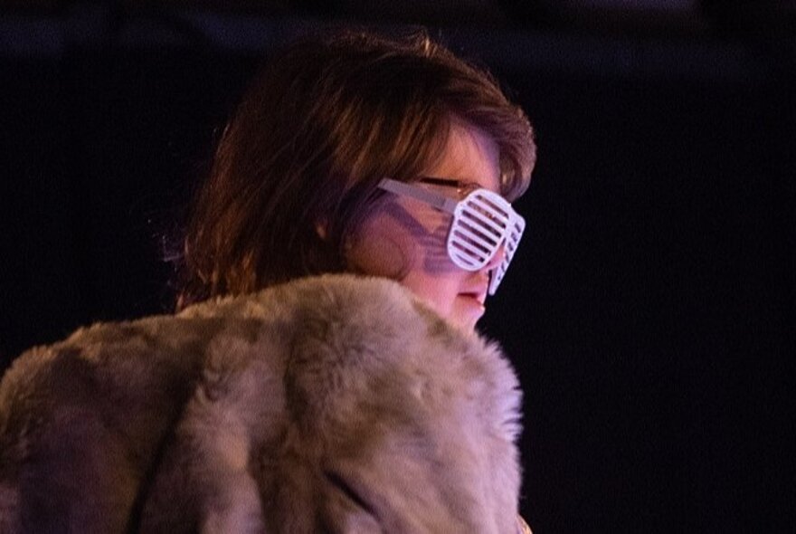 A woman wearing a light brown fur coat and striped shades, against a black background.