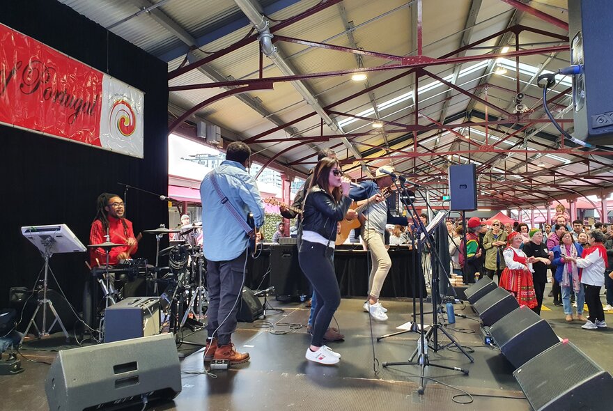 A band performing on a stage in a shed at Queen Victoria Market as part of the Taste of Portugal festival, with onlookers.