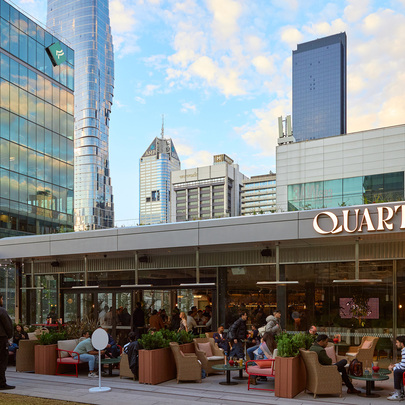 A crowd of people are seated outside a modern-style pub with a view of the city skyline behind them. There is a large sign saying Quarterhouse.