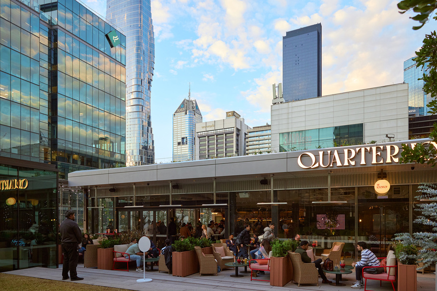 A crowd of people are seated outside a modern-style pub with a view of the city skyline behind them. There is a large sign saying Quarterhouse.
