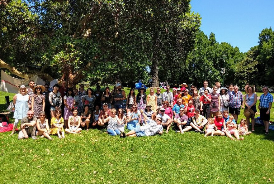 Large group of people sitting and standing on a grassy lawn underneath a large tree.