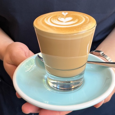 A glass cup of latte coffee with latte art, served on a light blue saucer and held by a person wearing a navy apron.