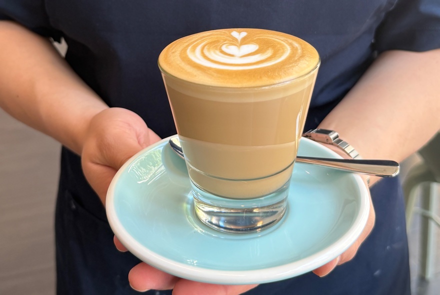 A glass cup of latte coffee with latte art, served on a light blue saucer and held by a person wearing a navy apron.