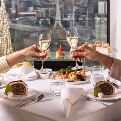 People toasting with raised champagne glasses over a formal lunch table with white linen, the Melbourne skyline in the background. 