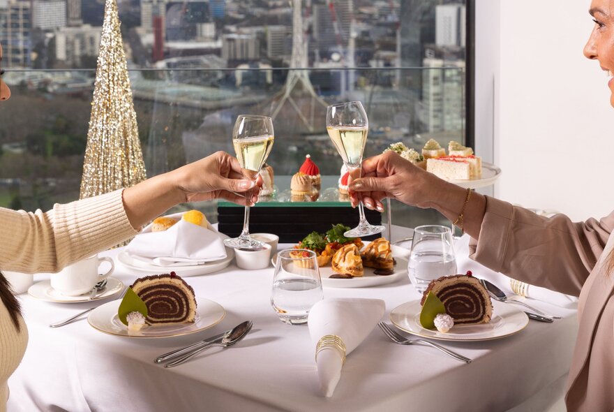 People toasting with raised champagne glasses over a formal lunch table with white linen, the Melbourne skyline in the background. 