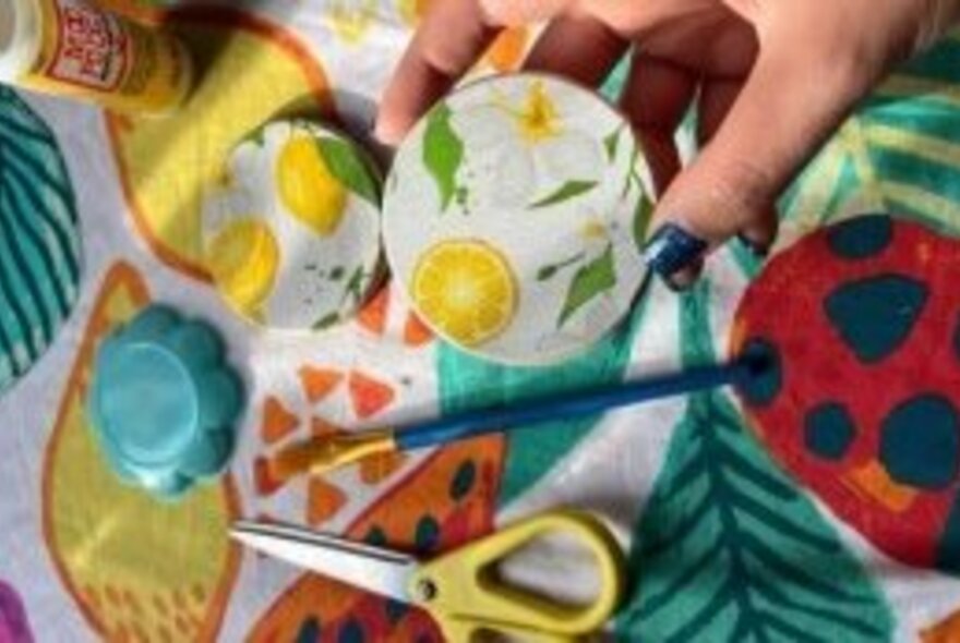 A hand holding a small coaster that has been decoupaged, with scissors and glue on the craft table in the background. 