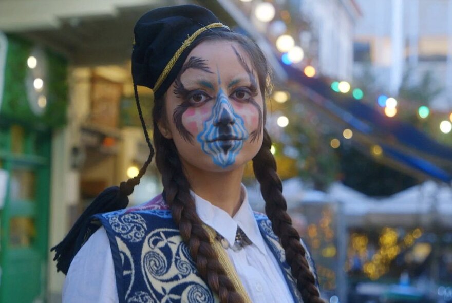 A person with a painted face and long plaits looking serious outdoors. 