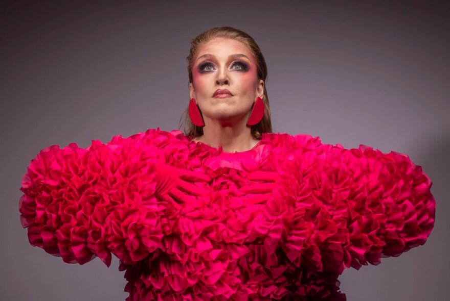 Performer wearing bright pink ruffled outfit with pink make-up and earrings, her arms raised in front of her.