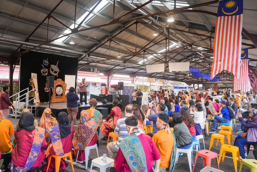 People seated in rows of chairs watching a performance on a small stage at the Colours of Malaysia festival at Queen Victoria Market.