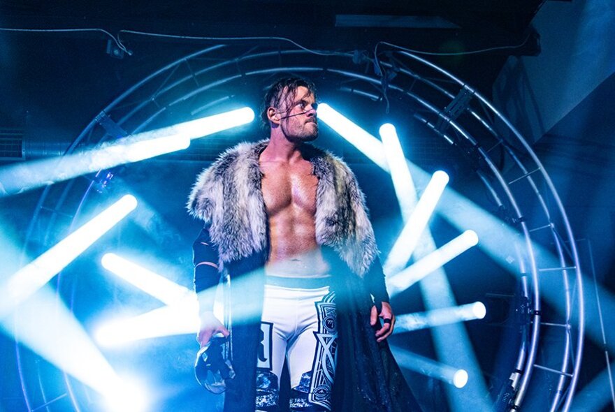 A wrestler with bare chest and a fur coat around his shoulders stands in front of illuminated signage. 