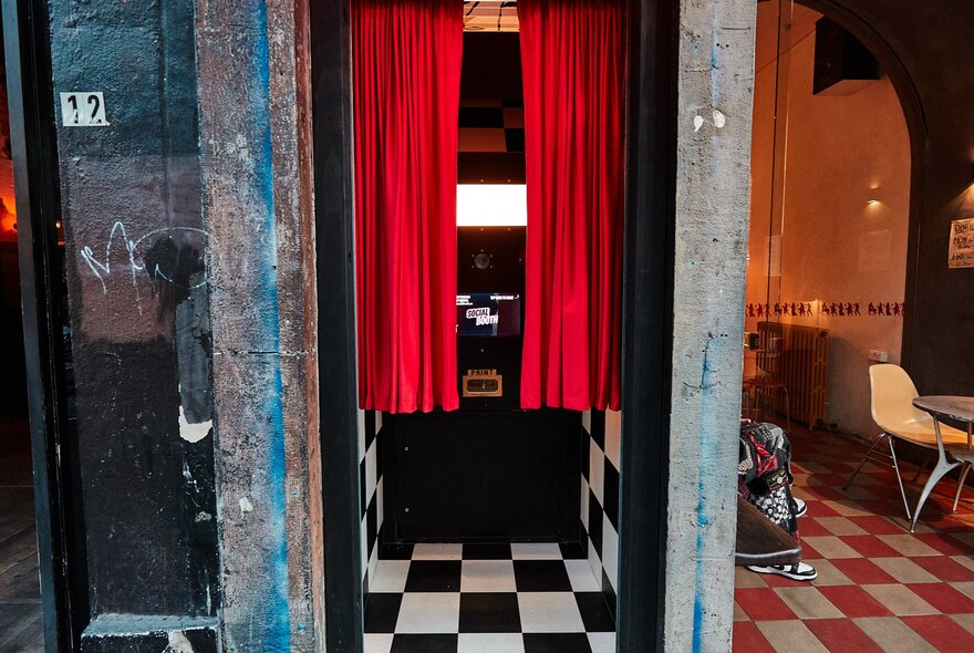 Vintage-style photo booth with red curtains and check floor, next to restaurant interior with similar design and arched windows.
