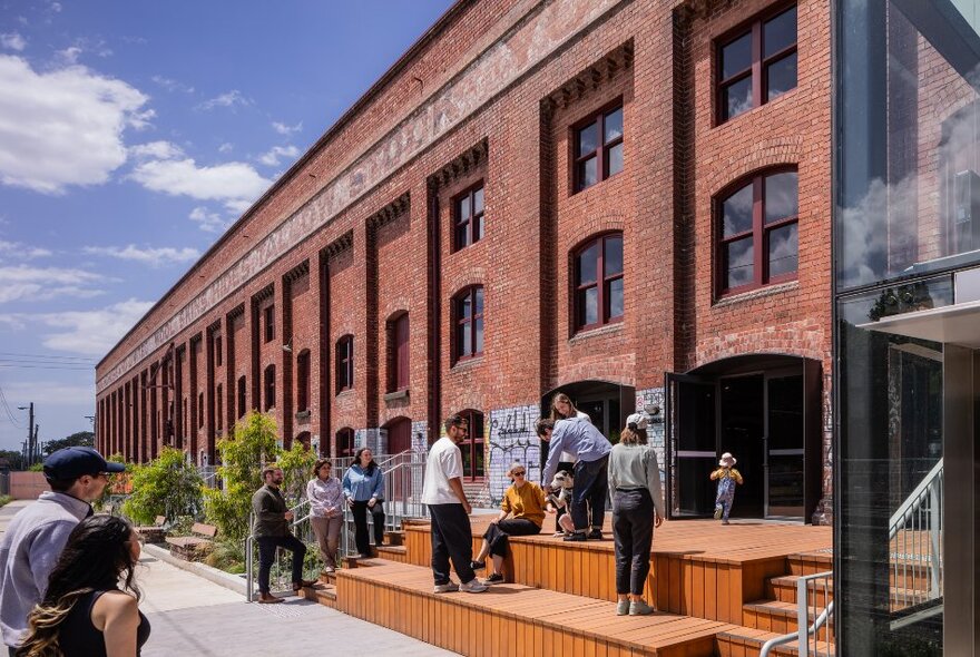 People on steps outside a redeveloped industrial precinct with red brick warehouse buildings.