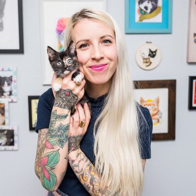 Author and educator Hannah Shaw, widely known as the Kitten Lady, holding a small kitten, her blond hair falling to one side of her face, she stands in front of a wall of cat pictures.