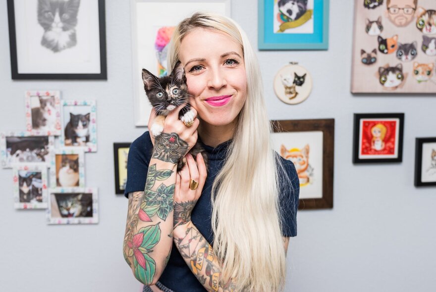 Author and educator Hannah Shaw, widely known as the Kitten Lady, holding a small kitten, her blond hair falling to one side of her face, she stands in front of a wall of cat pictures.