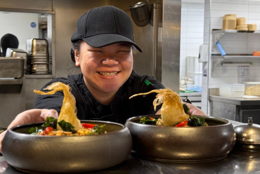 A chef in a black cap smiles as two dishes go out on the pass.