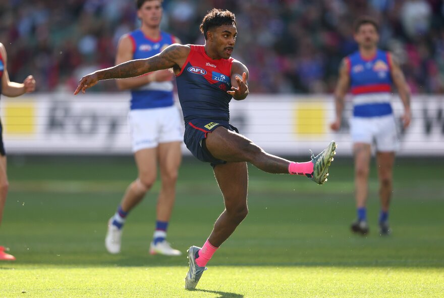 A Melbourne AFL football player kicking the football  during a match with two Western Bulldogs players behind him.