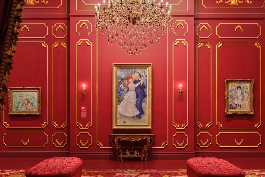 An art gallery with red walls and a chandelier hanging from the ceiling. There is a painting of two people dancing hanging on the wall. 